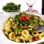 Pasta con rucola e pomodoro: un primo piatto sano e veloce 1 su tavolo con tovaglia bianca, un piatto bianco con pasta con rucola e pomodoro (orecchiette), vicino un bicchiere d'acqua e un contenitore di rucola