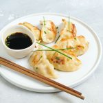 Ravioli cinesi al vapore: la ricetta dei jiaozi fatti in casa 1 In primo piano su un tavolo di marmo bianco c'è un piatto bianco fondo con dei ravioli cinesi. Accanto una piccola ciotola con la salsa di soia e due bacchette di legno.