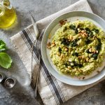 Risotto con zucchine: un primo piatto semplice da preparare 1 su tovaglia grigia, canovaccio con sopra un piatto che contiene risotto con zucchine e frutta secca con forchette e coltello, a sinistra basilico e contenitore di olio EVO