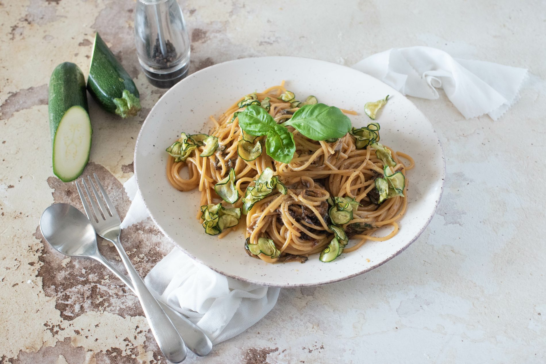 Sughi per pasta fatti in casa: ricette veloci e sane piene di gusto 3 su tavolo di marmo, piatto fondo con spaghetti alla Nerano con zucchine e basilico, sulla sinistra cucchiaio e forchetta e dietro il piatto una zucchina tagliata in 2 e un contenitore pepe