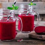 Succo di barbabietola: come prepararlo, benefici e proprietà 1 su tavolo di legno 2 bicchieri di vetro con cannuccia pieni di succo di barbabietola, a destra tagliere con fette di barbabietola