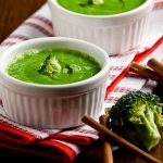 Vellutata di broccoli: una ricetta cremosa e dietetica 1 su tavolo di legno, canovaccio bianco e rosso piegato con 2 ciotole piccole con vellutata di broccoli con bacchette cinesi e accanto un pezzo di broccolo