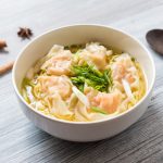 Wonton, la ricetta della pasta e del ripieno dei ravioli cinesi 1 su tavolo di legno ciotola con i wonton in brodo e verdure, sulla sinistra bacchette cinesi e a destra cucchiaio di legno