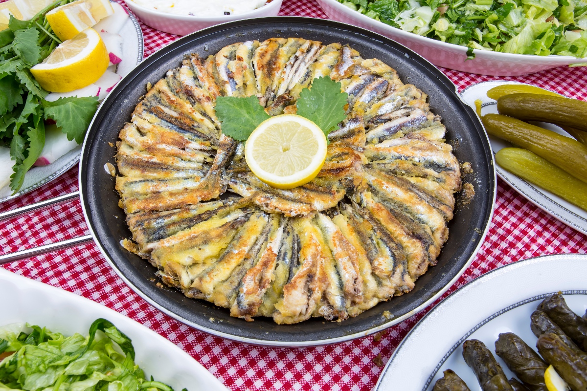 Ricette con alici: 12 idee semplici e ricche di sapore 4 su tavolo con una tovaglia a quadretti rossi e bianchi, piatti con insalate e alici in tortiera al centro del tavolo con un limone al centro