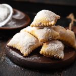 Bugie ripiene di Carnevale: la ricetta al forno fragrante 1 su tavolo di legno, tagliere con bugie ripiene di cioccolato, in fondo zucchero velato con un setaccio