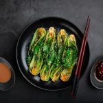 Pak choi in wok: ricetta leggera del cavolo cinese 1 su un tavolo nero, piatto scuro con cavolo cinese tagliato e bacchette cinesi. a destra e sinistra, ciotole con salse esotiche
