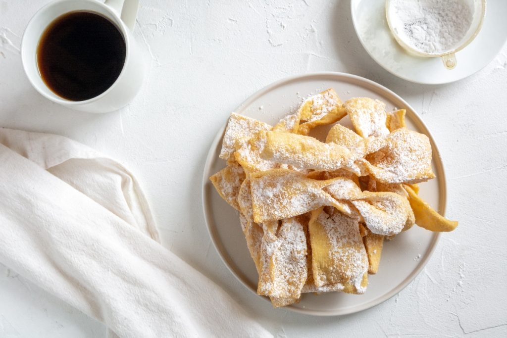 Dolci di Carnevale, ricette tradizionali e varianti al forno 1 tavolo bianco da colazione con chiacchiere senza glutine in un piatto bianco, sulla sinistra una tazza di caffè, accanto un tovagliolo bianco