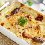 Parmigiana di patate perfetta: una ricetta cremosa, filante e facilissima da fare! 1 su tavolo bianco, tagliere di legno con piatto bianco di ceramica con parmigiana di patate. In alto a destra patate tagliate in 2