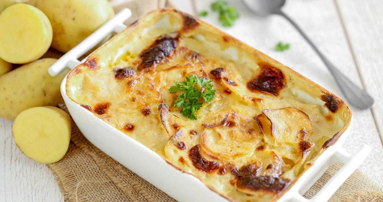 Ricette con le patate: le migliori da provare facilmente a casa 22 su tavolo bianco, tagliere di legno con piatto bianco di ceramica con parmigiana di patate. In alto a destra patate tagliate in 2