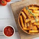 Polenta al forno: un primo piatto senza glutine per tutti 1 su tavolo bianco tagliere di legno con ciotola con la polenta al forno e parmigiano grattugiato e erbe, accanto piccola ciotola con salsa di pomodoro e dietro pomodorini