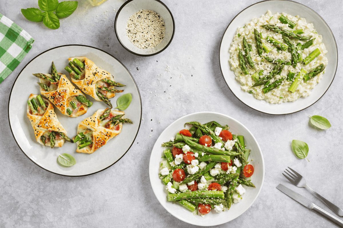 In primo piano su un tavolo in marmo chiaro ci sono dei piatti con alcune ricette con asparagi. A sinistra dei fagottini di pasta sfoglia con asparagi, in basso a destra un'insalata fresca con feta e pomodorini e in alto a sinistra un classico risotto con asparagi.