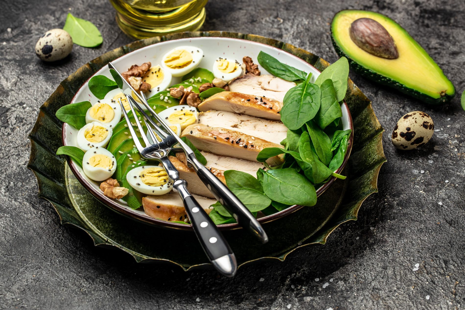 In primo piano su un piano di marmo scuro c'&egrave; un piatto con del pollo cotto alla piastra e delle uova sode e foglie di spinaci. Sullo sfondo un mezzo avocado e dell'olio evo.