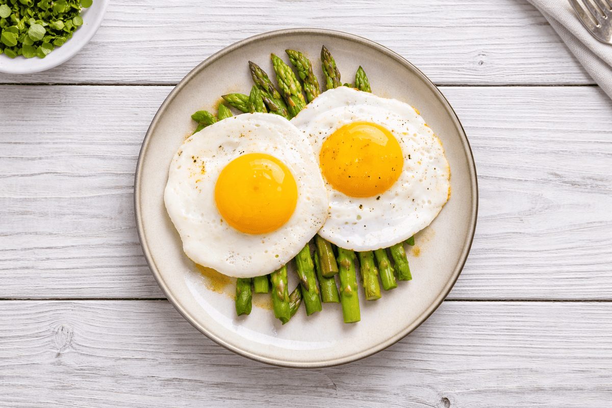 In primo piano su un tavolo di legno chiaro c'&egrave; un piatto fondo con degli asparagi alla milanese.