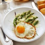 Asparagi alla milanese: un secondo di stagione facile e veloce 1 su una tavolo apparecchiata, piatto bianco con asparagi alla milanese con asparagi e uova con forchetta e coltello con tovaglioli bianchi , un calice di vino e panini