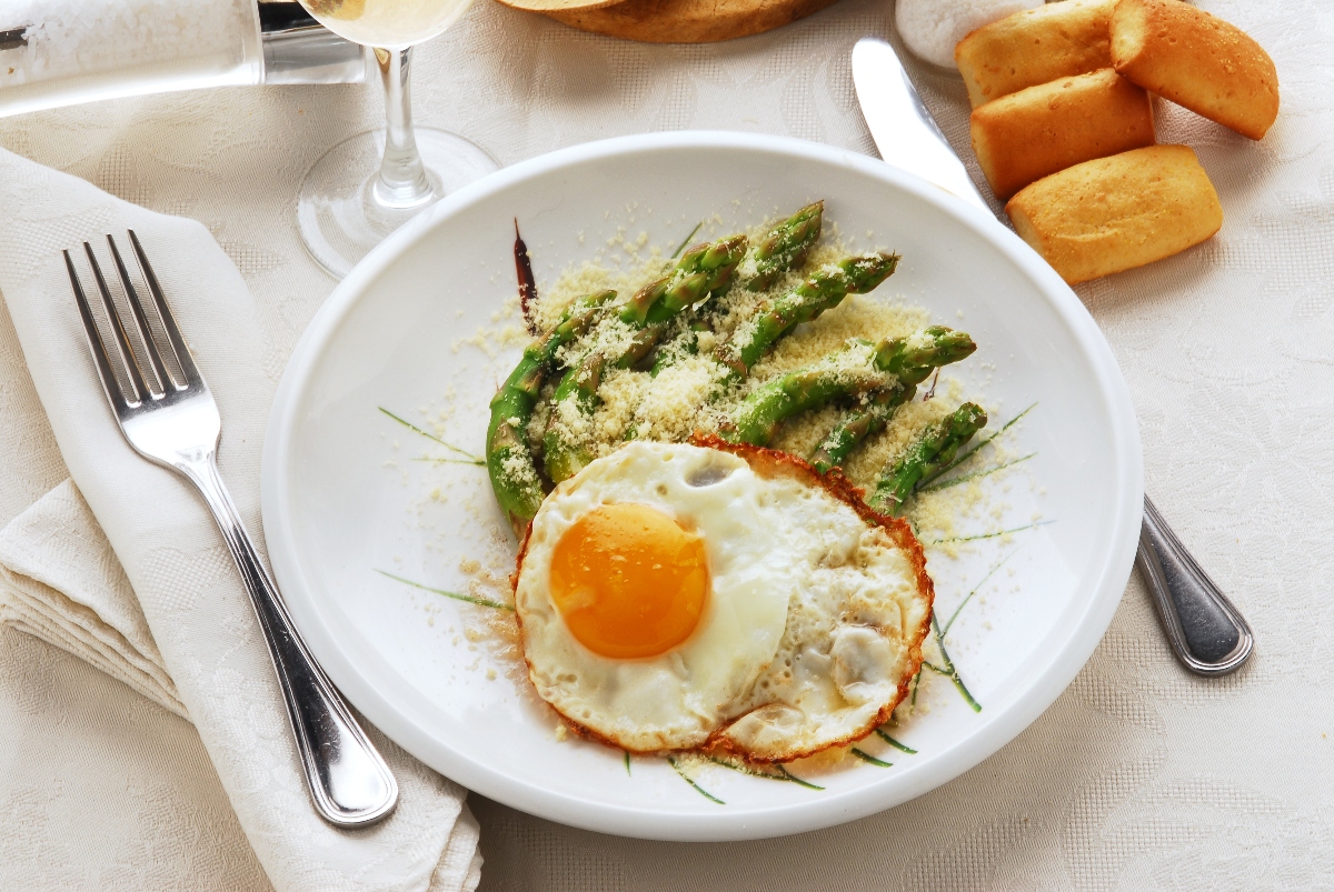 ricette con uova: su una tavolo apparecchiata, piatto bianco con asparagi alla milanese con asparagi e uova con forchetta e coltello con tovaglioli bianchi , un calice di vino e panini