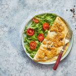 Frittata di albumi al forno: la ricetta con poche calorie 1 In primo piano su un tavolo di marmo grigio c'è un piatto fondo con una frittata di albumi e della rucola con i pomodorini, sul piatto c'è una forchetta con il manico in legno.