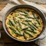 Frittata di asparagi, un secondo piatto primaverile 1 su tavolo di legno canovaccio beige con piatto da forno che contiene una frittata di asparagi ,sulla destra piattino con asparagi puliti