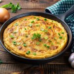 Frittata di patate e cipolle: una ricetta sfiziosa 1 ricette con frittata: su tavolo di legno, teglia con frittata di patate e cipolle con attorno, cipolle bianche e rosse, un canovaccio verde a quadretti bianchi