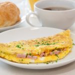 Omelette prosciutto e formaggio 1 su un tavolo da colazione in primo piano piatto bianco con omelette prosciutto e formaggio, dietro una tazza di caffè e accanto una brioche in un piatto bianco