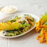 Pasta con fiori di zucca: un primo piatto delicato e gustoso 1 ricette con fiori di zucca: su tavolo di legno bianco, piatto piccolo con la pasta e sopra un fiore di zucca. Accanto 3 fiori e dietro una ciotola con parmigiano grattugiato