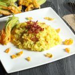 Risotto con fiori di zucca: un primo piatto delizioso 1 su tavolo di legno scuro, piatto bianco quadrato con risotto con fiori di zucca e all'angolo fiore di zucca per decorare, anche dietro e sulla destra forchetta e coltello