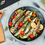 Sardine al forno: una ricetta light e saporita 1 su piano cottura, piatto nero con sardine al forno con pomodorini e limoni a fette, a destra tagliere con coltello e sulla sinistra rosmarino