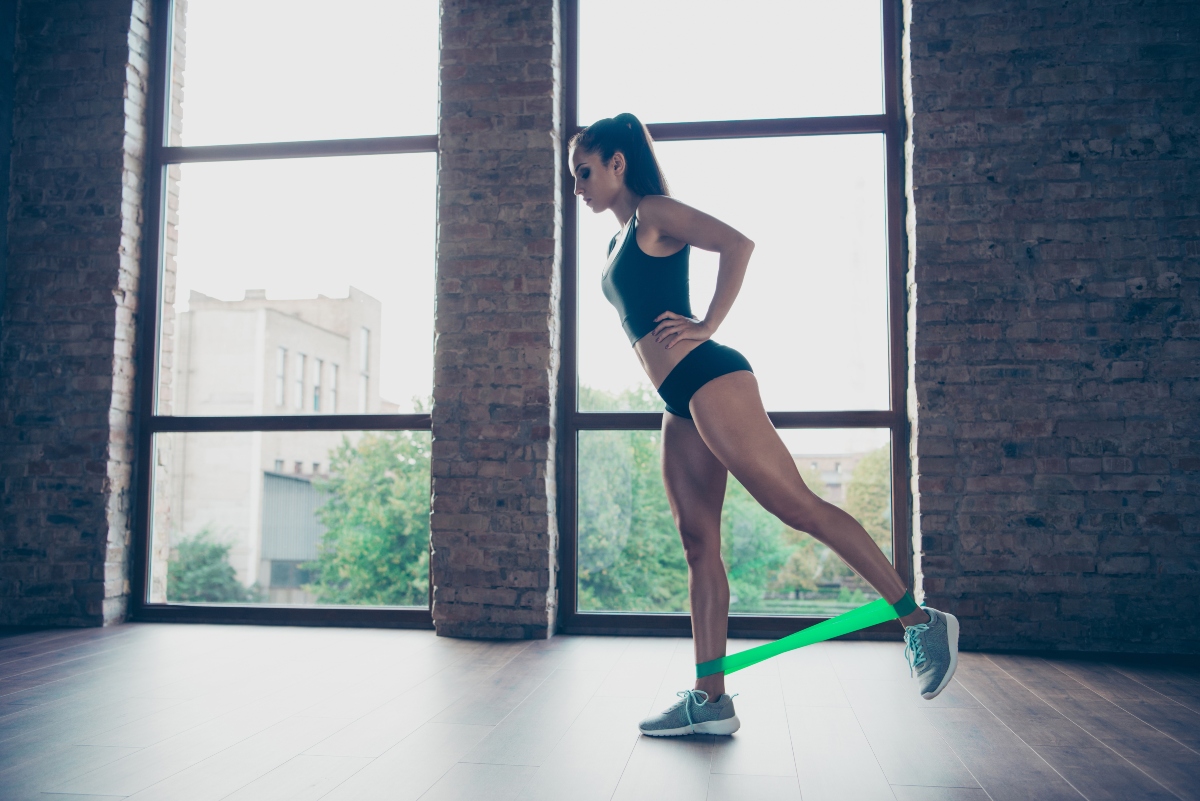 Sedere piatto: cause, rimedi ed esercizi per un sedere tonico e voluminoso 1 Donna in casa vestita da shorts e top esegue slanci in piedi con elastico fitness per combattere il sedere piatto