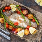 Sogliola al forno: un secondo piatto leggero e gustoso 1 su tavolo di legno sotto tavolo stile bohémien con un piatto con sogliola al forno, patate, asparagi, pomodorini con alcune fette di limoni . Sulla sinistra forchetta e coltello