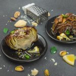 Carciofi bolliti: un contorno veloce, sano e gustoso 1 su tavolo di marmo, 2 piatti neri con carciofi bolliti, attorno pistacchi, fette di limone, grattugia e parmigiano reggiano