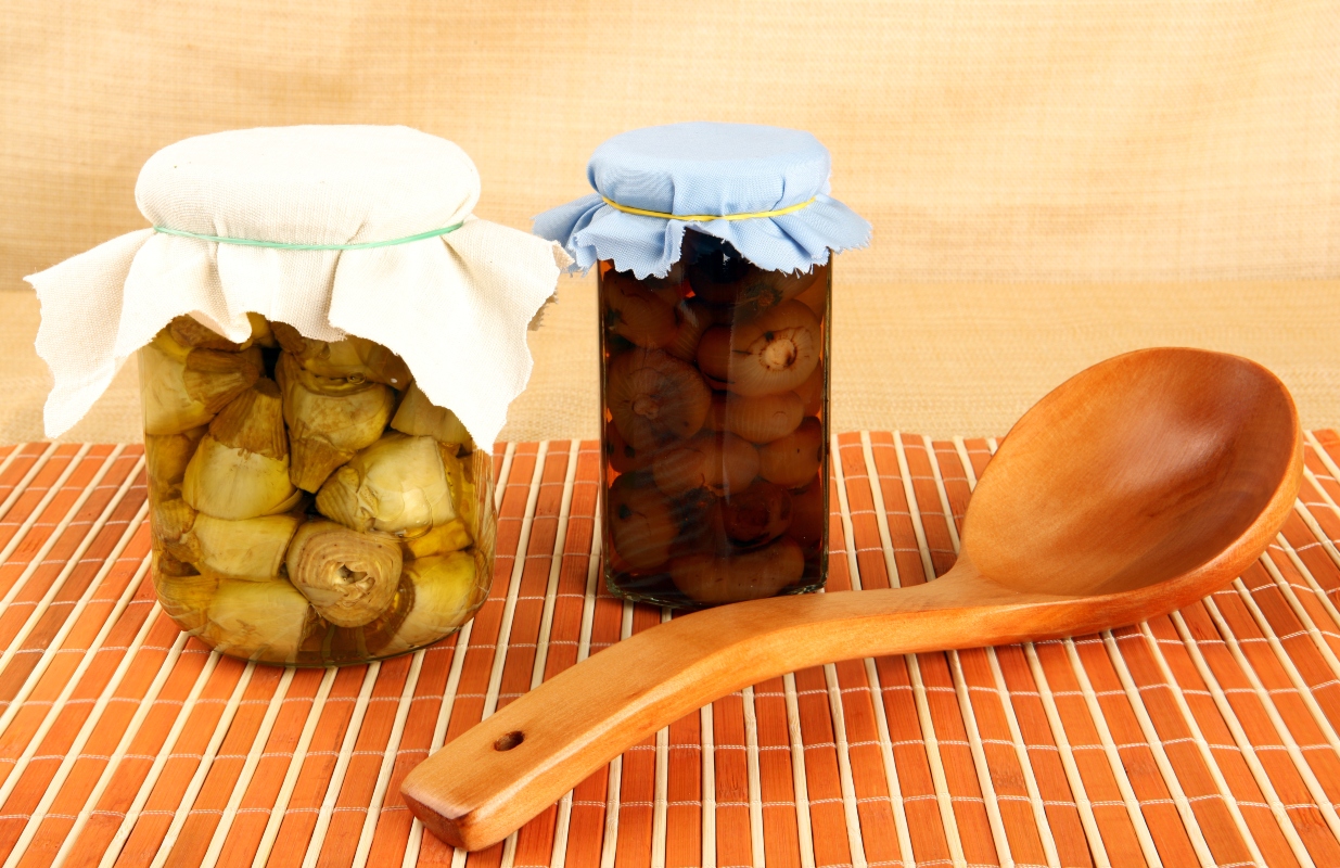 Carciofini sott'olio fatti in casa: una ricetta deliziosa 2 su tavolo di cucina con tovaglia di vimini arancione con strisce beige con sopra 2 conserve fatte i casa: carciofini sott'olio e cipollino sott'olio. A destra cucchiaio di legno