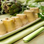 Carciofini sott'olio fatti in casa: una ricetta deliziosa 1 su tavolo da cucina piatto rettangolare verde sopra una tovagliolo verde con 6 carciofini sott'olio e sulla destra una forchetta verde. dietro il piatto carciofi freschi