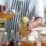 Come fare kombucha in casa: scopri la ricetta del tè nero fermentato 1 come fare kombucha: uomo in cucina con grembiule che sta preparando kombucha in casa e riempie i bicchieri