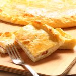Parigina: la ricetta della pizza rustica napoletana 1 primo piano di un tagliere di legno con una parigina, pizza napoletana ripiena di prosciutto e pomodori con 2 fette tagliate e una forchetta