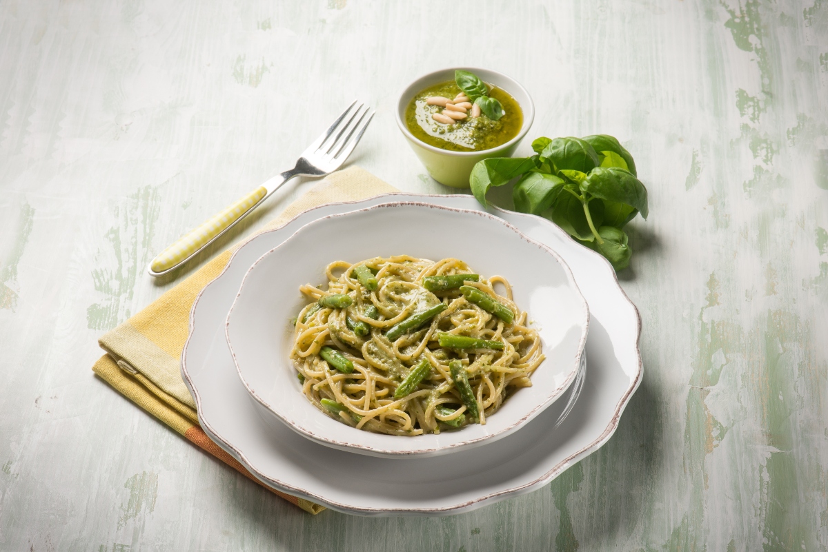 Ricette con fagiolini: 11 idee facili, gustose e nutrienti 4 su tavolo bianco apparecchiato piatto bianco con pasta con fagiolini dietro basilico e accanto forchetta gialla e tovagliolo giallo