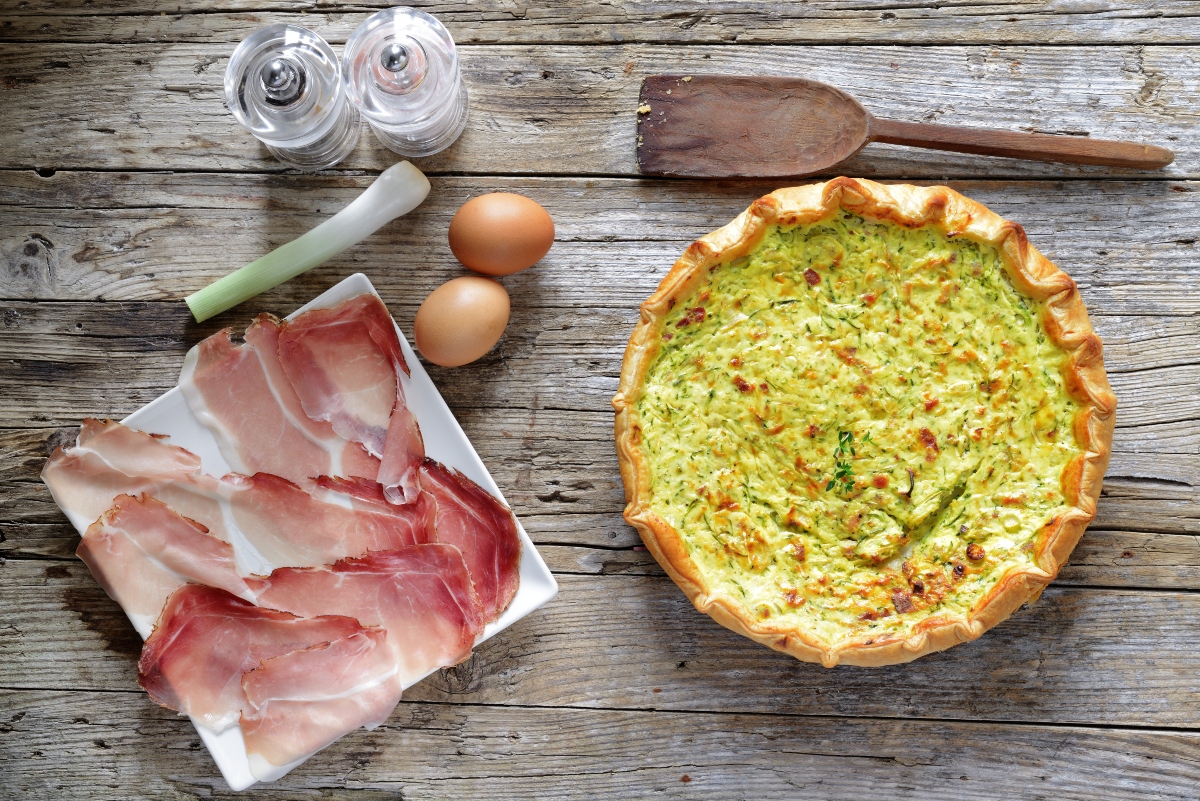 Torte salate: 29 ricette veloci e leggere per tutti i gusti 7 torta salata zucchina e speck su una tavola di legno, sulla sinistra piatto bianco con fette di speck e accanto 2 uova e un pezzo di porro, saliera e pepiera e dietro un cucchiaio di legno