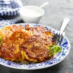 Rosti di patate: le golose fritelle cotte al forno 1 Su tavolo di marmo, piatto bianco con fiori blu con rosti di patate e sotto insalata verde, sul bordo del piatto 1 forchetta e dietro ciotola piccolo con salsa di yogurt e un canovaccio