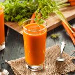 Succo di carota fatto in casa: benefici e ricetta con estrattore e Bimby 1 su tavolo di legno, bicchiere con succo di carota e prezzemolo, dietro tagliere con mazzo di carote