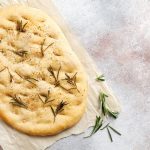 Focaccia: ricetta base per un impasto perfetto 1 in primo piano su della carta forno una focaccia bianca con sale grosso e rametti di rosmarino