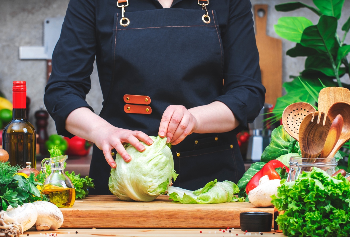 Insalata iceberg: proprietà, benefici e come usarla in cucina 2 cuoca con grembiule nero davanti a piano di cucina che pulisce un'insalata iceberg