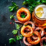 Polpo fritto: una ricetta leggera e facilissima 1 su tavolo di marmo, tagliere di marmo con un polpo fritto tagliato a rondelle con attorno erbe aromatiche e un bicchiere di tè al limone
