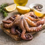 Polpo in pentola a pressione: ricetta e tempi di cottura 1 polpo in pentola a pressione: su tavolo tagliere con un polo in pentola a pressione, erbe aromatiche e limone tagliato, dietro una bottiglietta di olio EVO