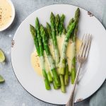 Salsa olandese senza burro: ricetta facile e veloce in 5 minuti 1 su piano da cucina di marmo piatto bianco con asparagi e salsa olandese accanto limone tagliato e ciotola con salsa