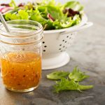 Vinaigrette: ricetta classica e le varianti 1 su tavolo ciotola con vinaigrette con frusta, dietro scolapasta in metallo bianco con insalata appena lavata
