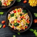Pasta fredda con tonno: piatto estivo nutriente 1 su sfondo nero ciotola nera con pasta fredda con tonno e mais e pomodori. Attorno foglie di basilico e pomodorini tagliati