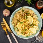 Pesto di limoni: sapore agrumato per una pasta leggera e dietetica 1 su tavolo di marmo, piatto azzurro con pasta con pesto di limoni e limoni tagliati a fette , accanto forchetto e coltello, limoni e grattugia, olio EVO e erbe aromatiche