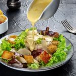 Salsa Caesar: la ricetta originale con ingredienti semplici e varianti light 1 in primo piano su un tavolo scuro c'è un piatto di caesar salad che viene condito dalla salsa caesar e crostini croccanti