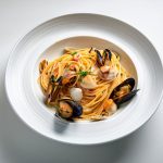 Spaghetti allo scoglio: primo piatto di mare con ingredienti e varianti 1 In primo piano su uno sfondo bianco c'è un piatto di spaghetti allo scoglio con frutti di mare come vongole e cozze.