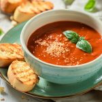 Zuppa di pomodoro cremosa: con pomodori o passata, da servire anche fredda 1 in primo piano un piatto piano color verde acqua con due crostoni di pane su cui poggia una ciotola con della zuppa di pomodoro decorata con una foglia di basilico.