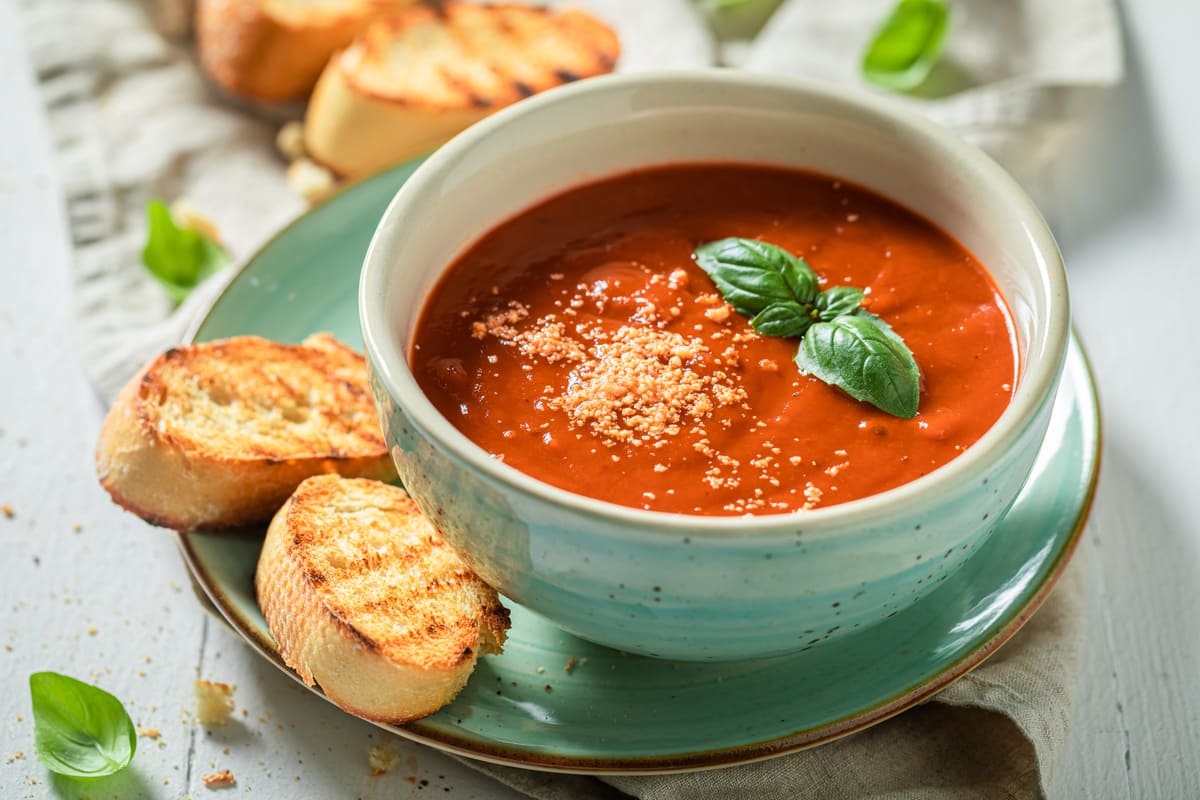 in primo piano un piatto piano color verde acqua con due crostoni di pane su cui poggia una ciotola con della zuppa di pomodoro decorata con una foglia di basilico.