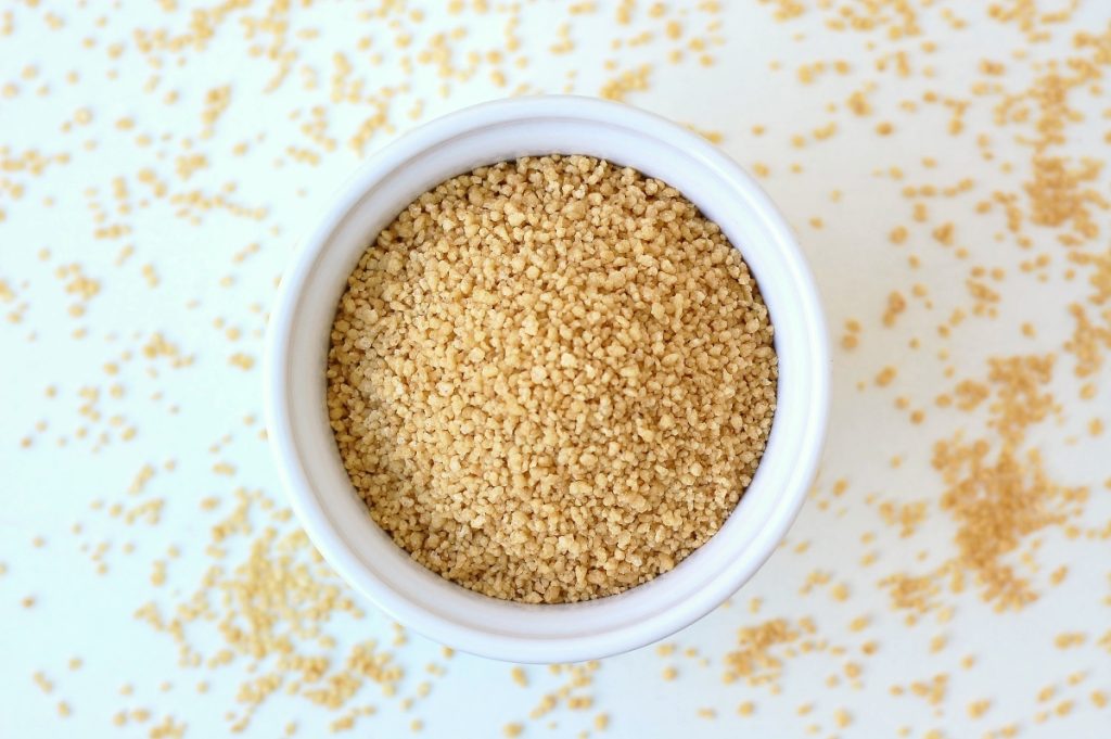 Cous cous integrale: calorie, benefici e ricette sane e gustose 1 su tavolo bianco, primo piano di una ciottola bianca con cous cous integrale e sul tavolo chicchi di cous cous sparsi